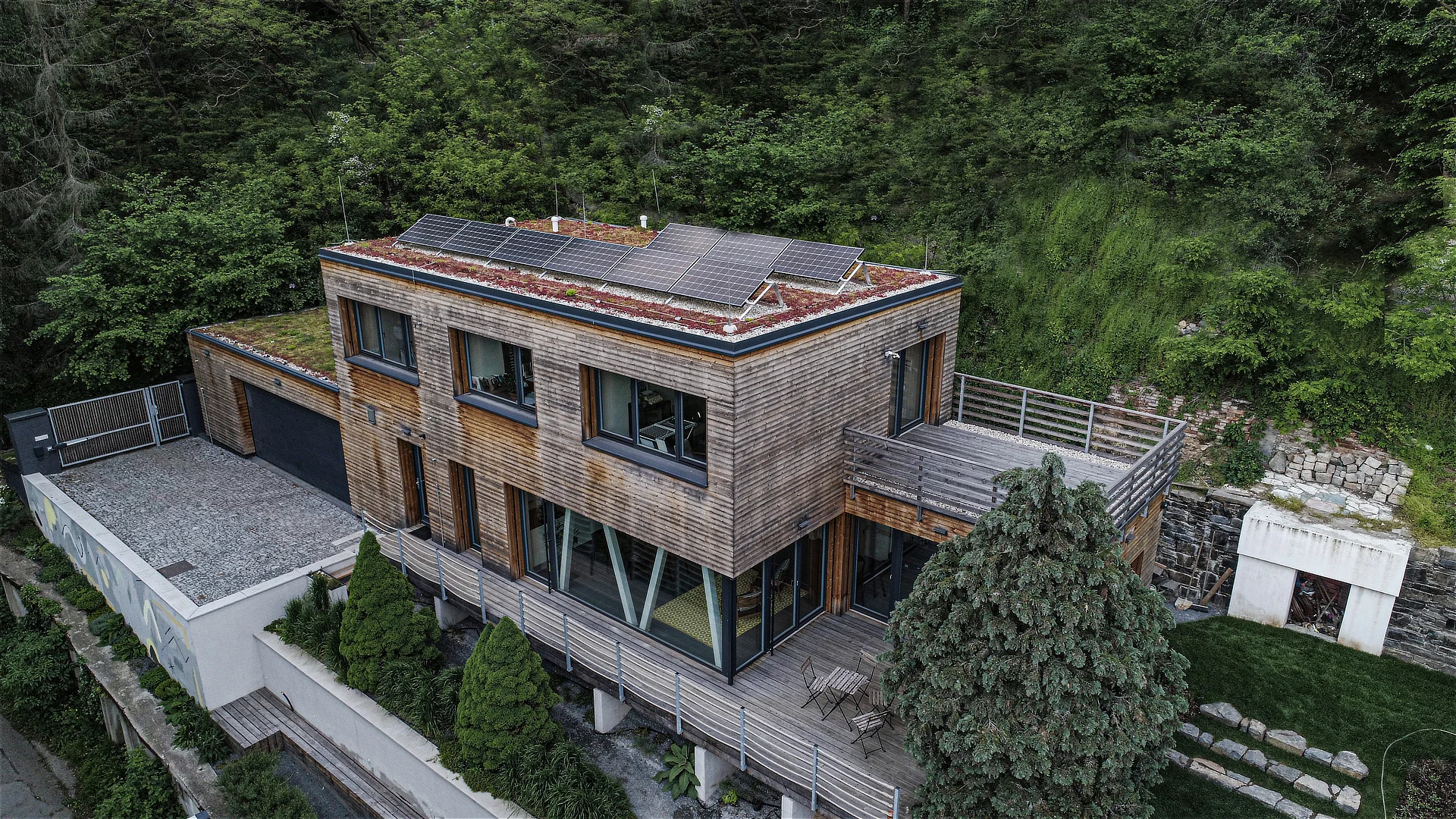 Nulový dům Zbraslav z masivních CLT panelů zasazený ve svahu, s dřevěnou fasádou, terasami a fotovoltaickými panely.