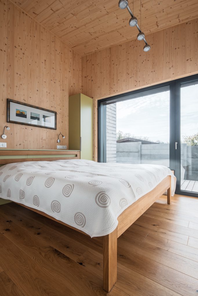 Ložnice s přiznanými CLT stěnami a velkým posuvným oknem vedoucím na terasu