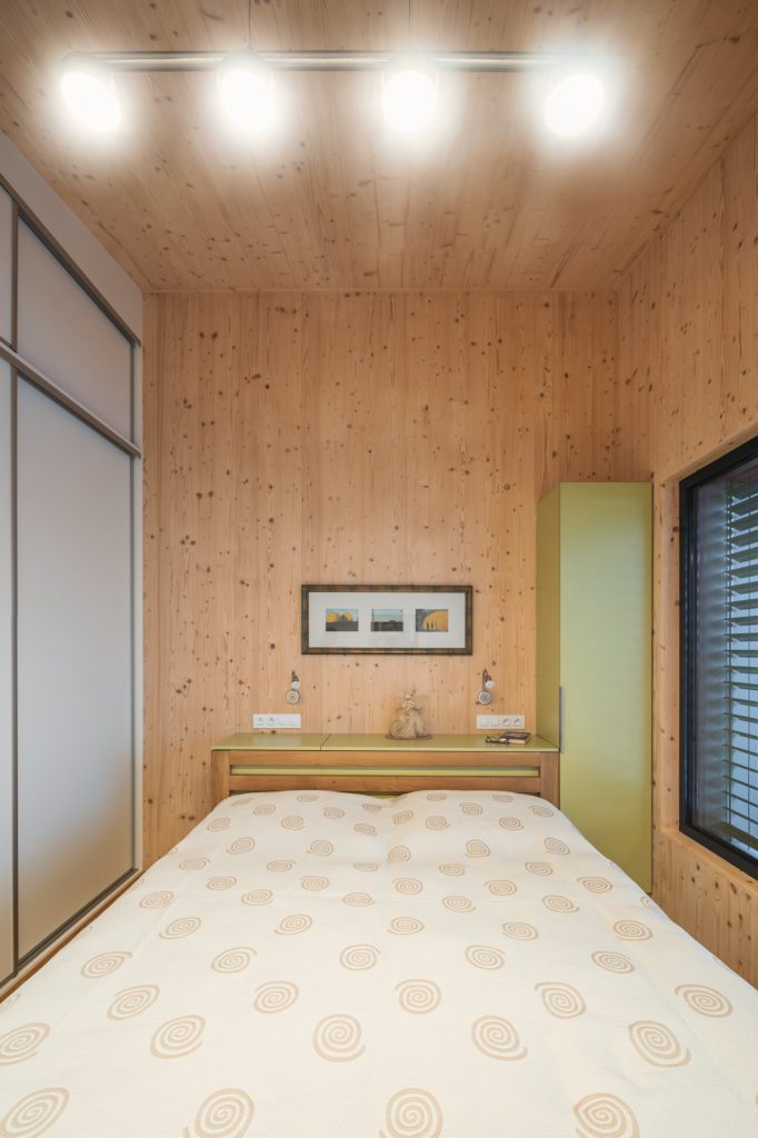 Ložnice s dřevěným obkladem z CLT panelů, vestavěnou skříní a oknem s venkovní žaluzií