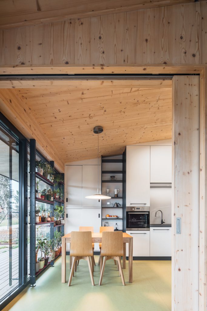 Bílá kuchyňská linka pod přiznaným dřevěným CLT stropem s velkým prosklením směrem do zahrady
