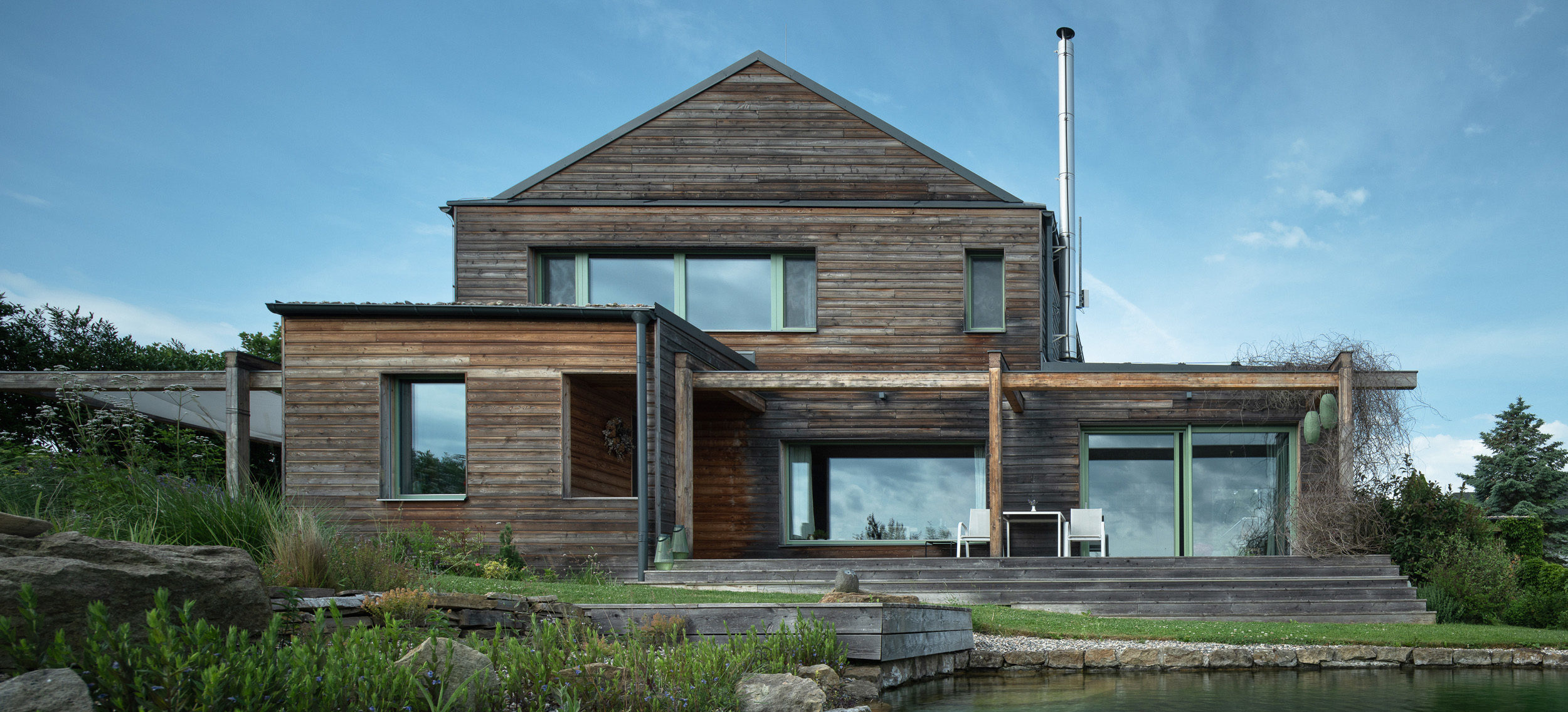 Dům pro dva z CLT panelů od firmy 3AE s dřevěnou fasádou a terasou u jezírka