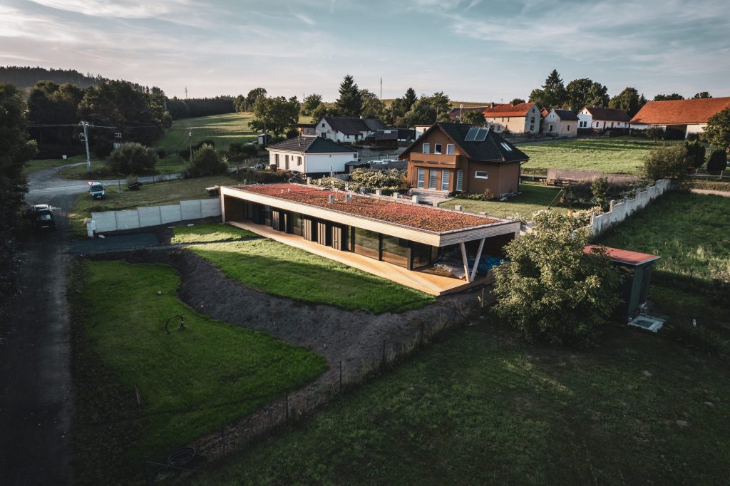 Přízemní dřevostavba s vegetační střechou a dřevěným obkladem na okraji vesnice