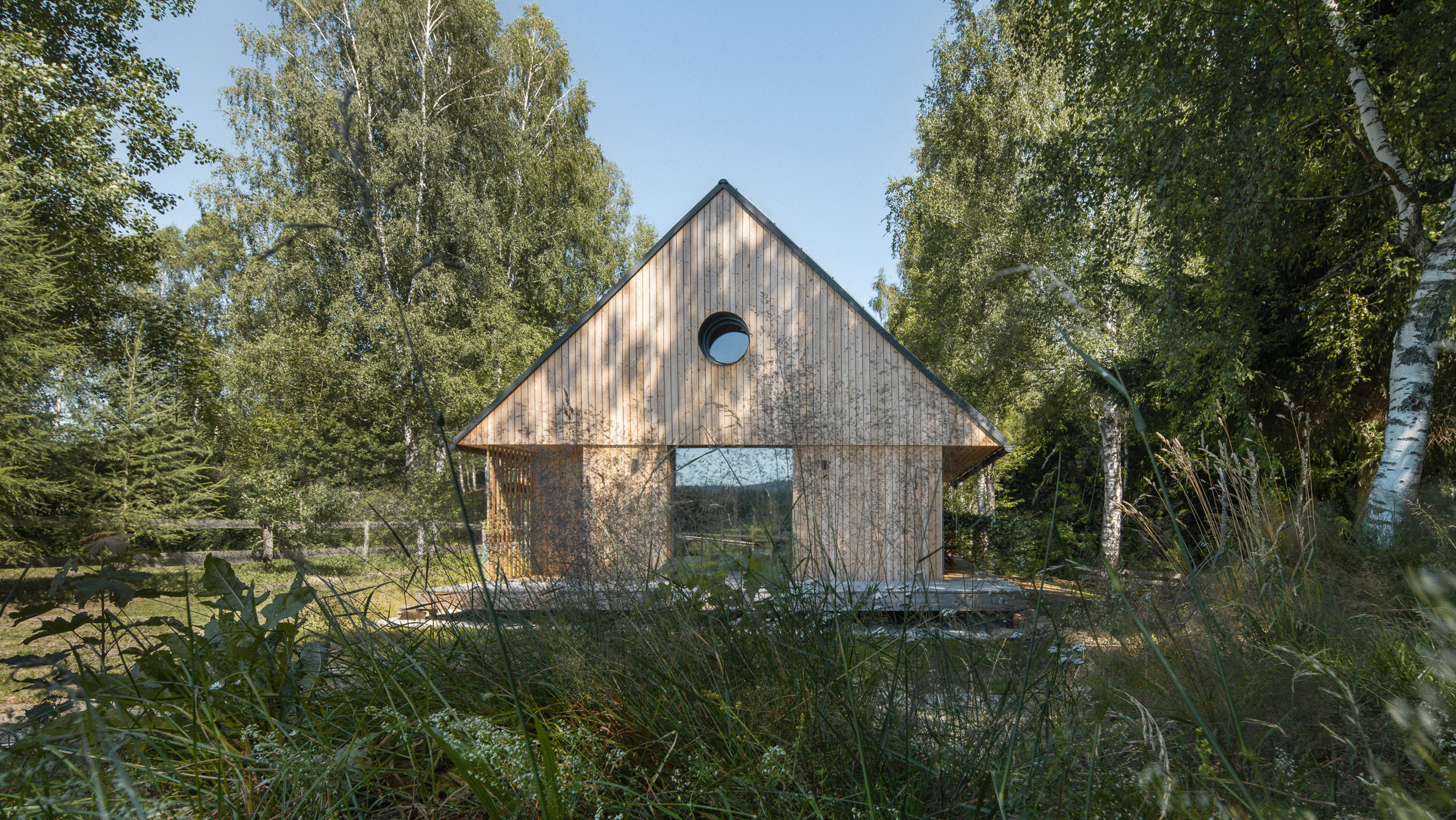 Dřevěná chata realizovaná firmou 3AE se sedlovou střechou a kulatým oknem mezi stromy