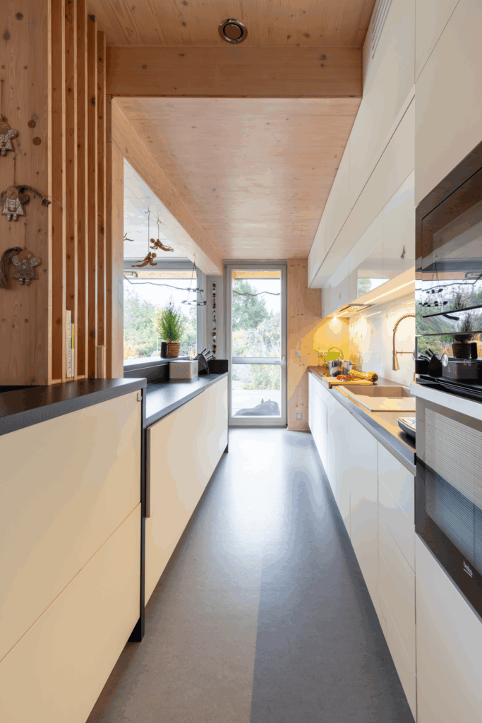 Úzká kuchyň s vestavěnými spotřebiči, bílým nábytkem a stropem z CLT panelů