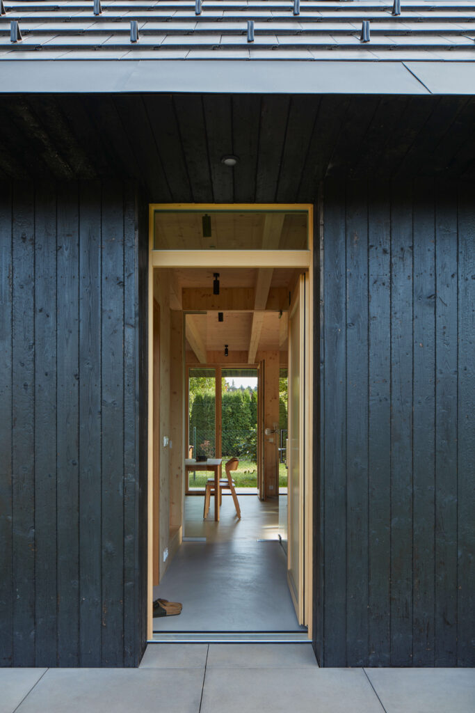 Vstupní část domu s opálenou dřevěnou fasádou a velkými okny.