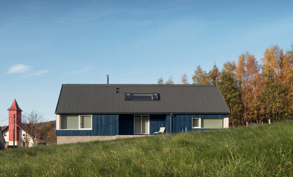 Chata s modrou fasádou, sedlovou střechou a soklem z pohledového betonu