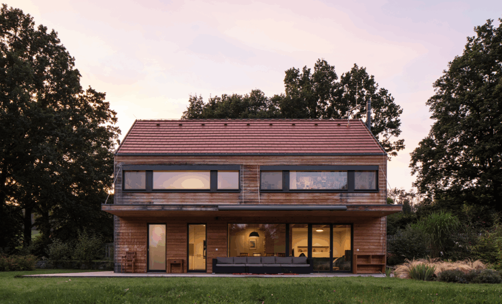 Dům se sedlovou střechou, dřevěnou fasádou a velkými okny při západu slunce