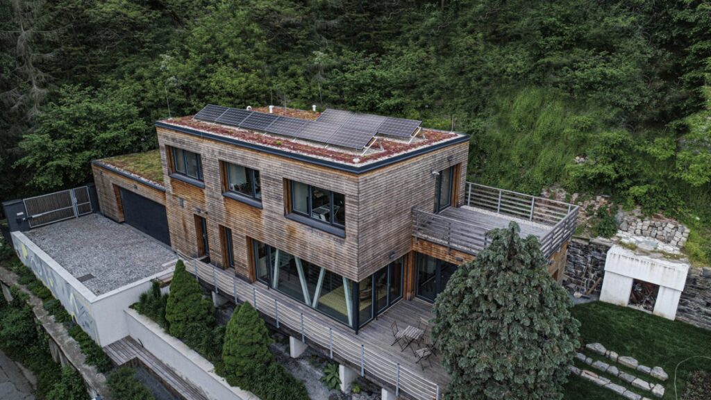 Dvoupodlažní dřevostavba ve svahu s dřevěnou fasádou, zelenou střechou a solárními panely
