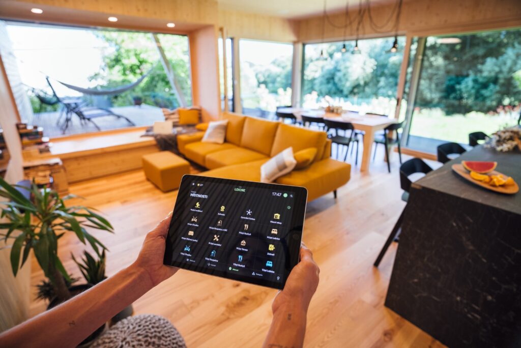 Tablet se systémem Loxone pro chytré ovládání domu v obývacím prostoru se žlutou sedačkou a výhledem na terasu.