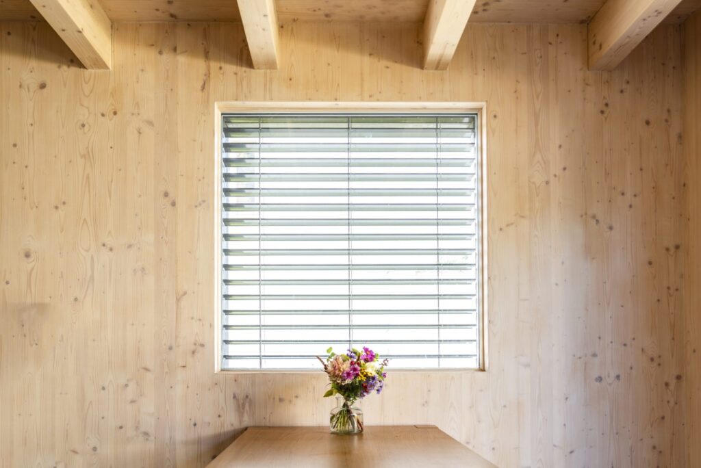 Dřevěný interiér s pohledovým CLT panelem, oknem se žaluzií a vázou s květinami