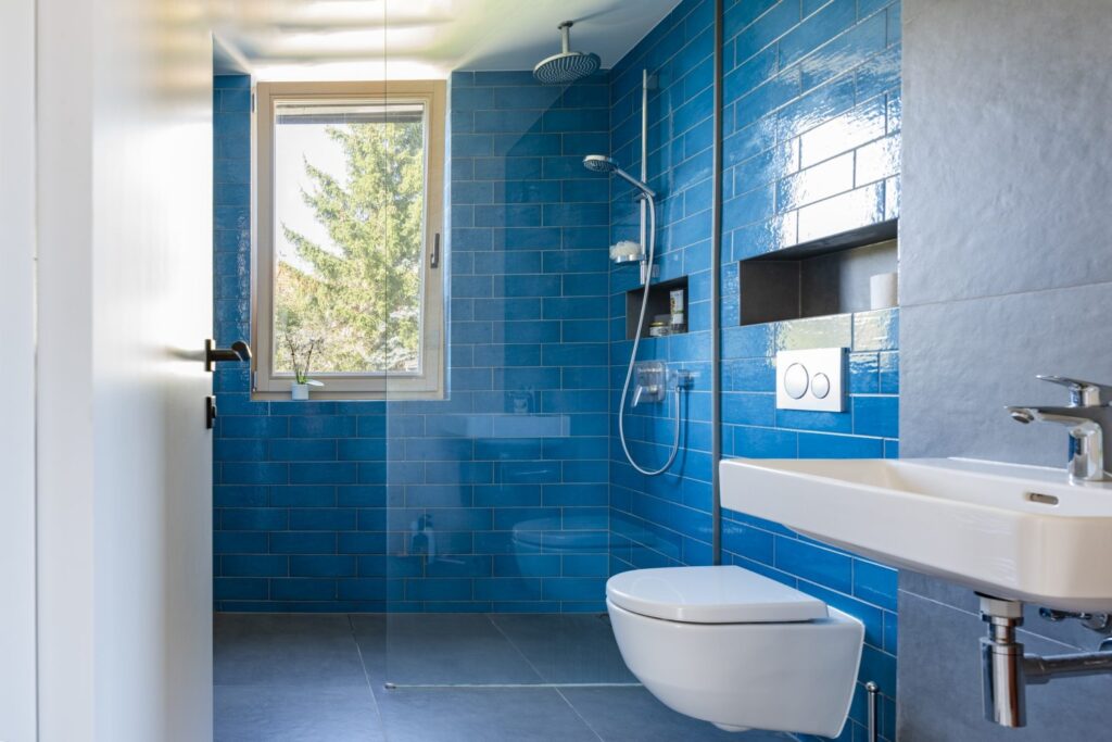 Koupelna s modrými obklady, sprchovým koutem, toaletou a oknem s výhledem na strom