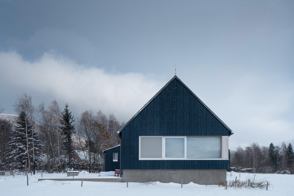 Modrá čelní fasáda chaty s velkým oknem a betonovým soklem ve sněhu