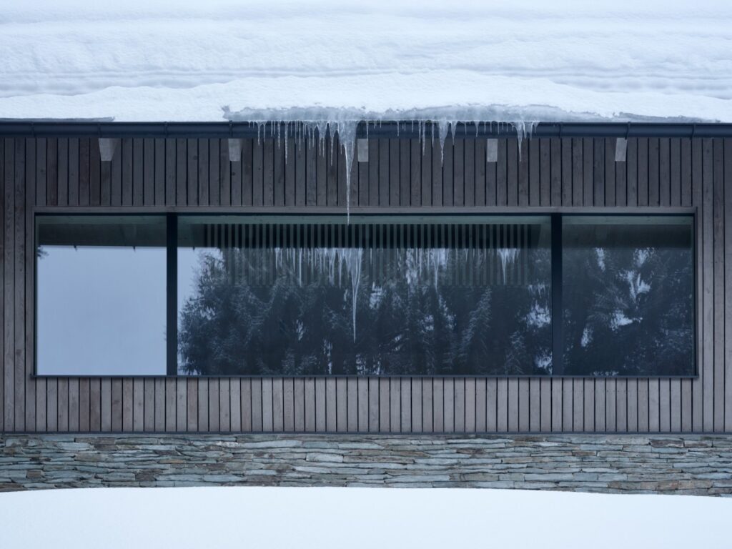 Detail velkého okna v dřevěné fasádě s rampouchy a kamenným soklem pod sněhem
