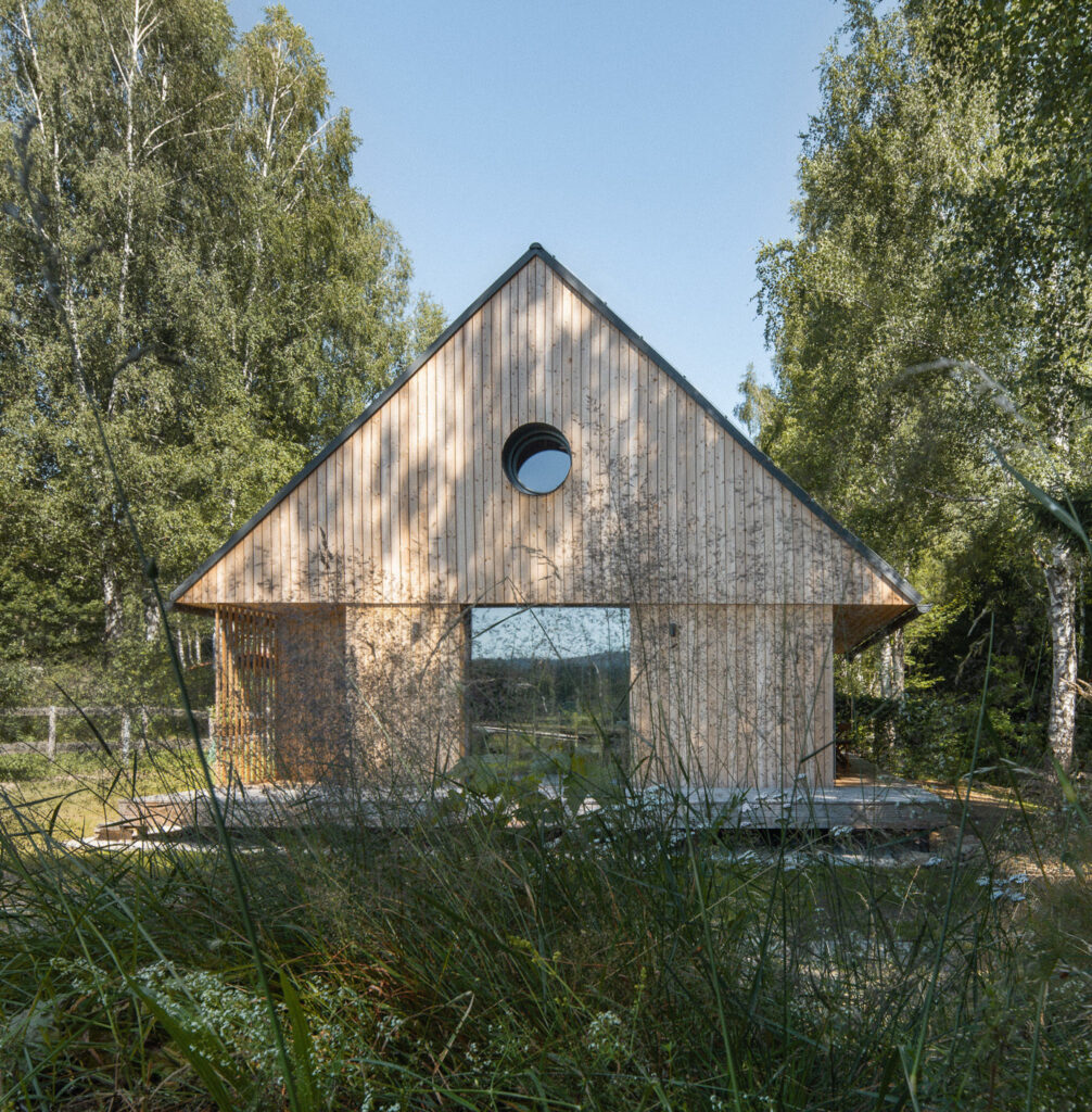 Dřevostavba s přírodní fasádou, sedlovou střechou a kruhovým oknem v lese