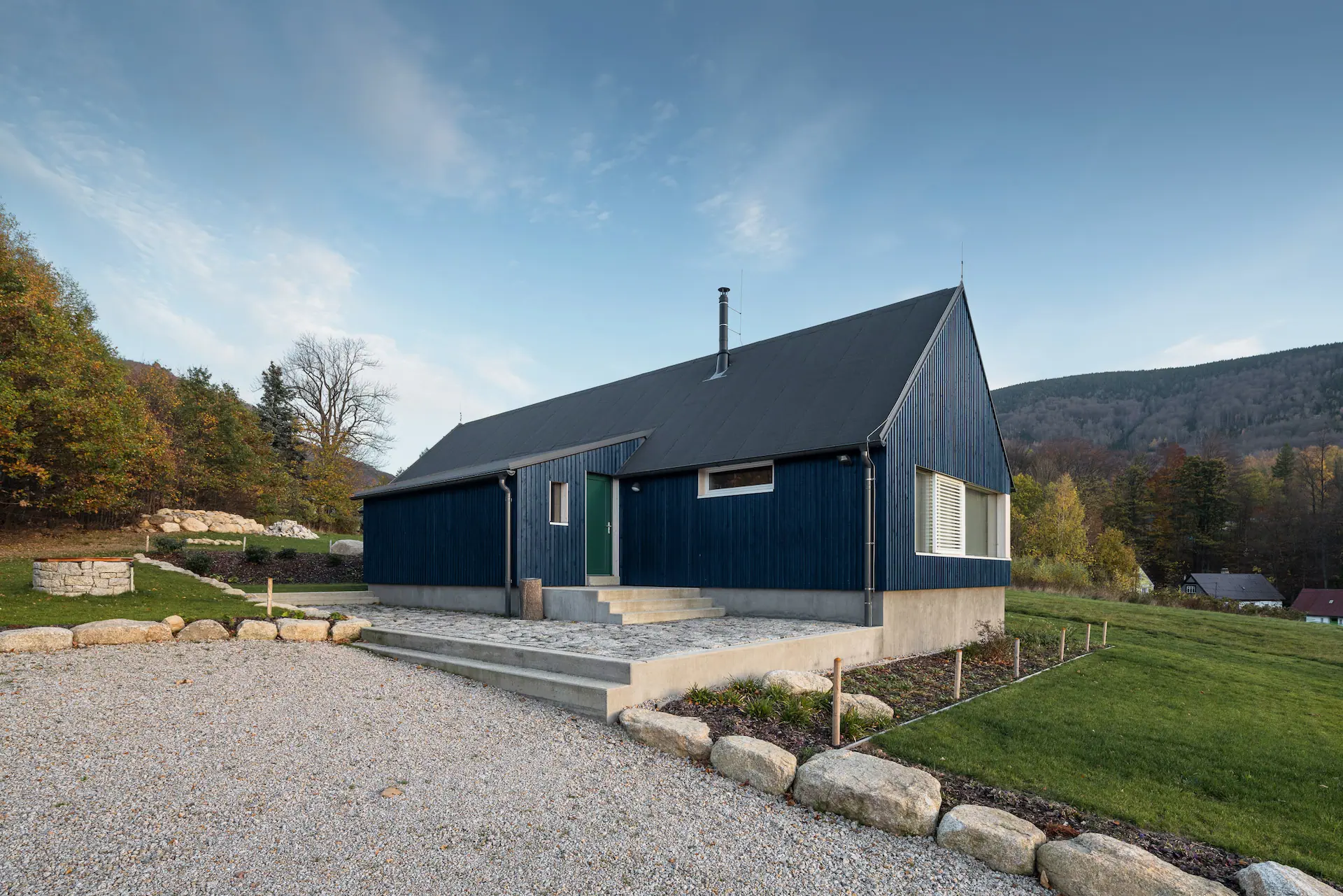 Vstupní část chaty s modrou dřevěnou fasádou, betonovým soklem a kamenným lemováním.