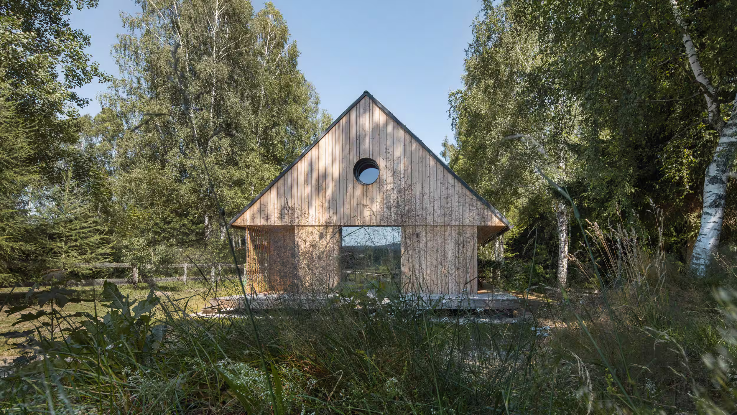 Dřevěná chata realizovaná firmou 3AE se sedlovou střechou a kulatým oknem mezi stromy