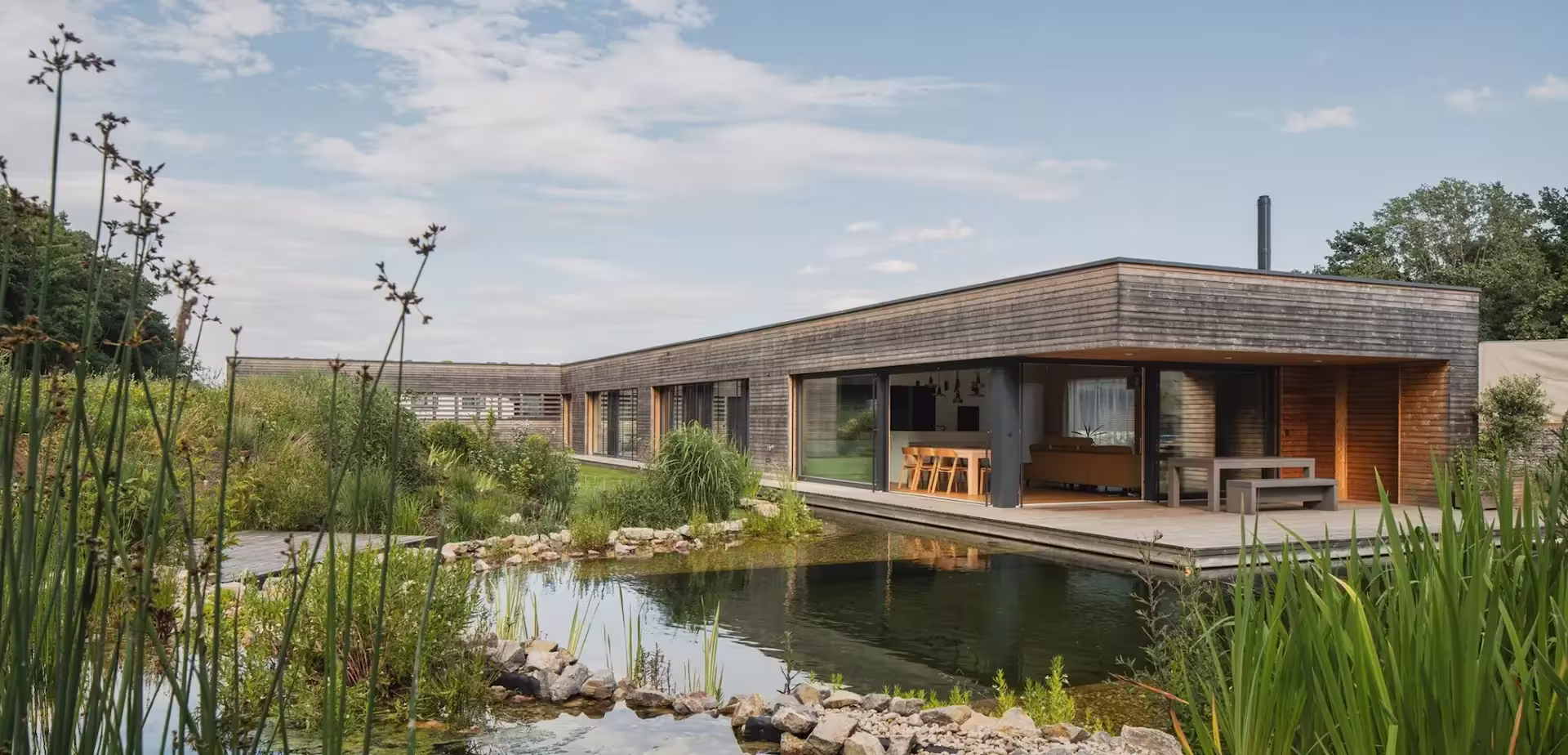 CLT dřevostavba ve tvaru L s terasou a přírodním jezírkem v zahradě