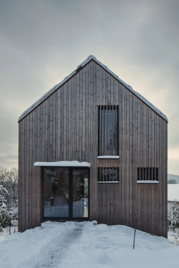 Čelní pohled na minimalistickou dřevostavbu v Plzni Liticích podle návrhu architekta Lukáše Janouta z Luni architekti se zasněženým pozemkem.