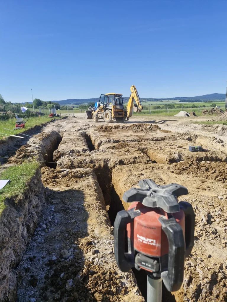 Aktivní výkopové práce základových pasů s laserovou nivelací Hilti v popředí na staveništi v Zalánech.